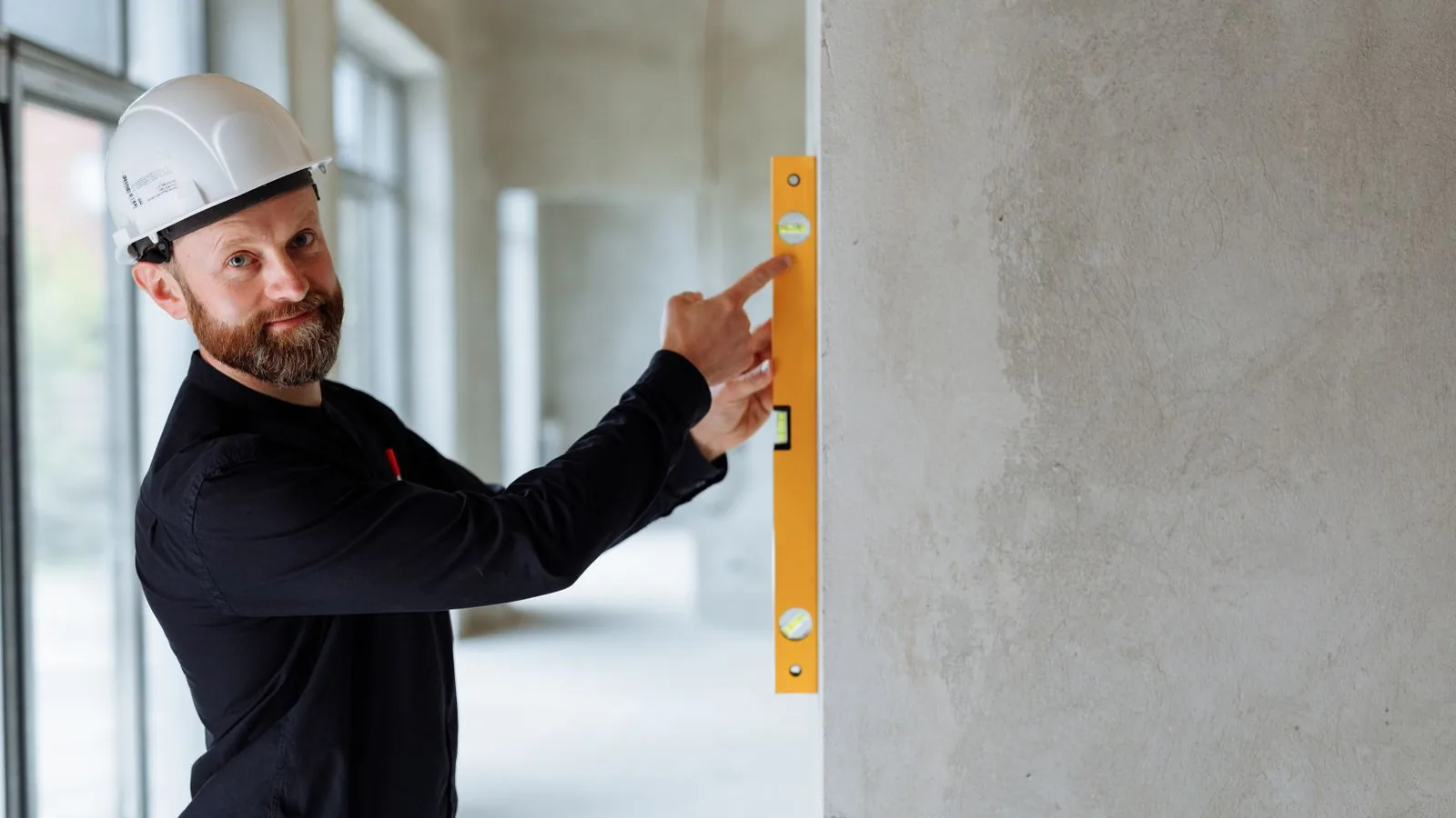 A site inspector in a hard hat holding a spirit level against a plastered wall to check it is plumb